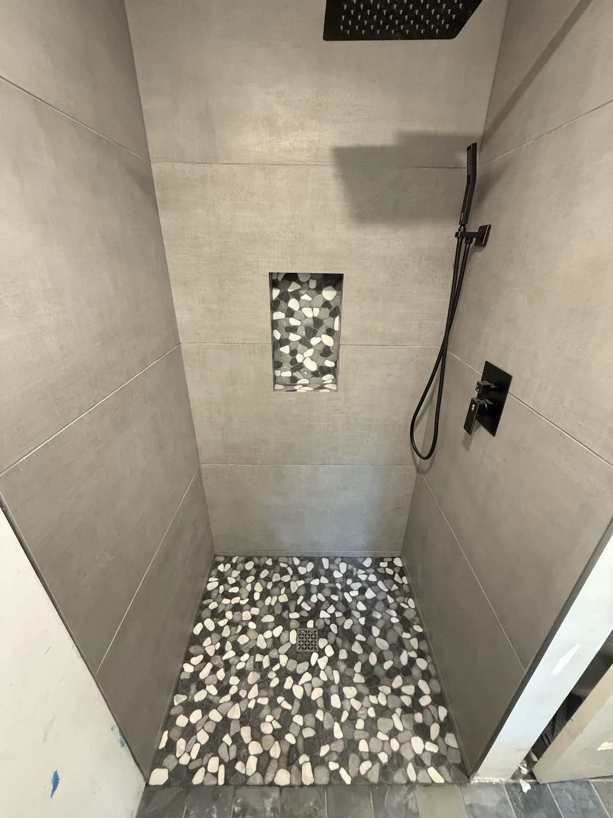 Complete custom tiled shower: large-format gray tile walls, pebble mosaic floor, niche, and matte black fixtures