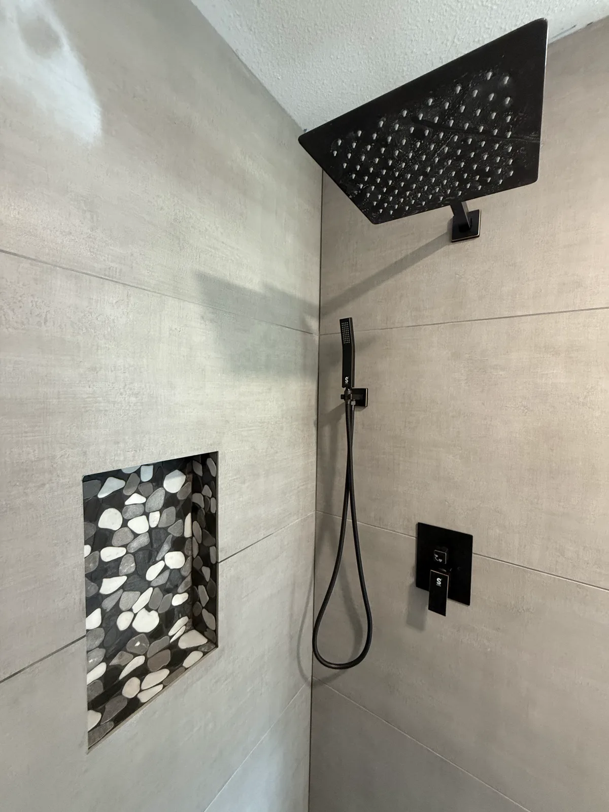 Close detail of matte black rain head, hand shower, valve, and pebble niche in a finished custom shower
